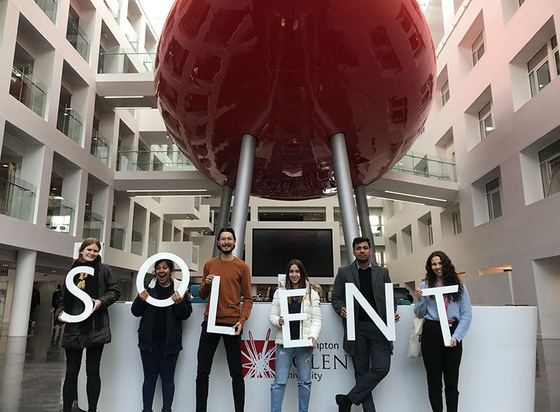 Solent's team of international alumni ambassadors standing in front of the pod holding a letters to spell Solent