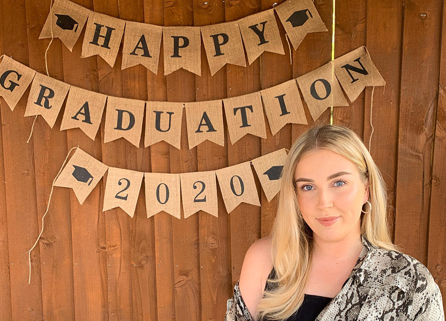 Picture showing Emily in front of banner that reads 'Happy graduation 2020'