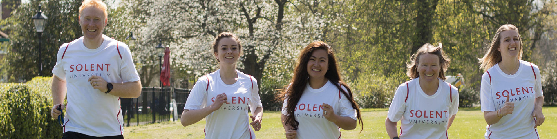 Five people run towards the camera across a park wearing Solent University white t-shirts.