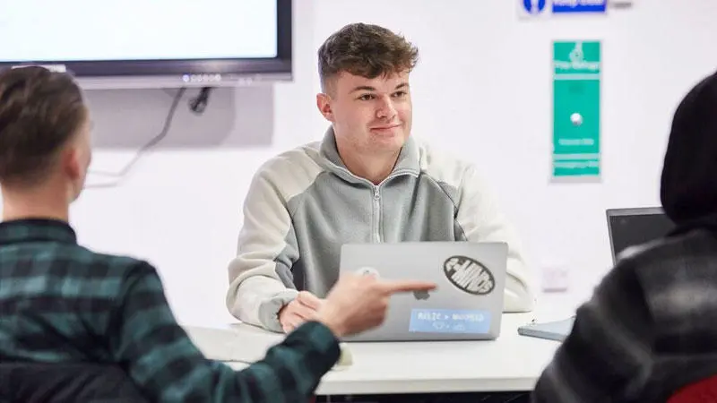 male student sat at desk with laptop in front of other students 