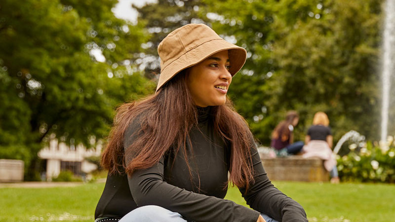 Two female students sat on the grass in the park with a fountain in the background