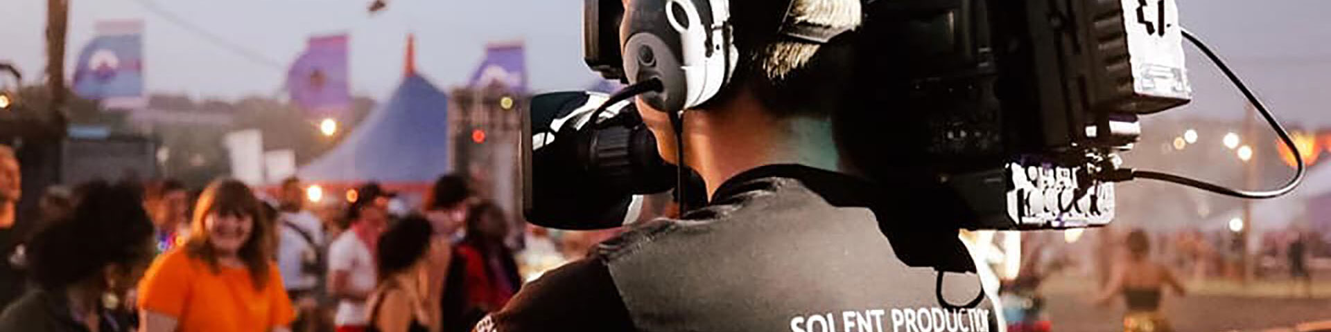 A man points a camera on his shoulder at some people at a music festival.