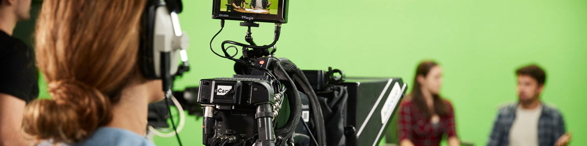 A student with headphones on looks down a large TV camera at a boy and girl sat down on two chairs behind a table and in front of a green screen.