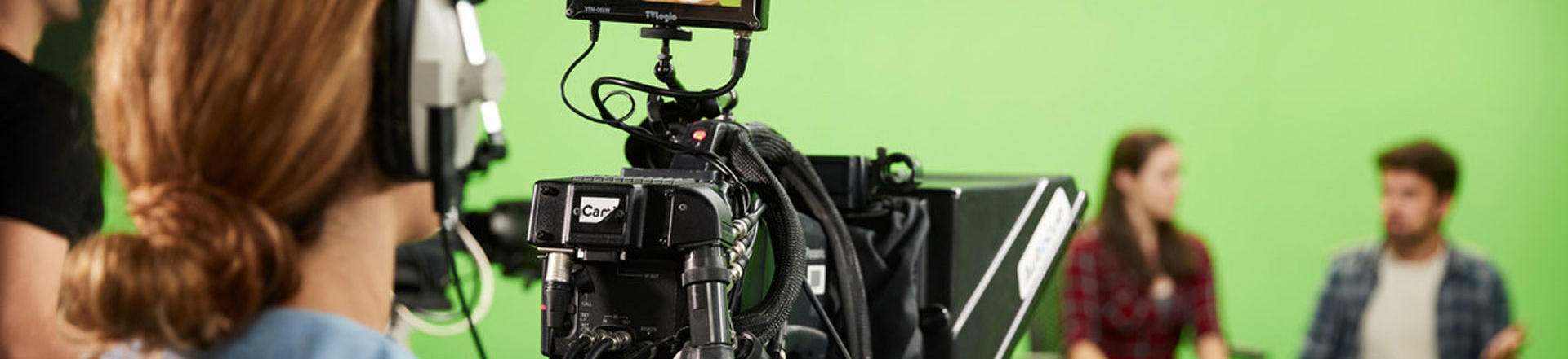 A student looks down a TV camera at two students sat at a desk in front of a green screen.