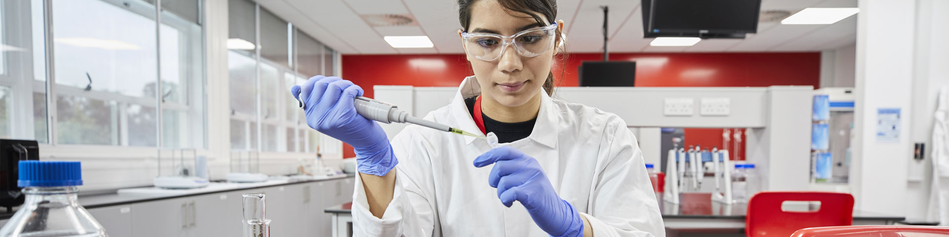 A woman in a lab coat wearing blue gloves and sat down in a lab fills a test tube.