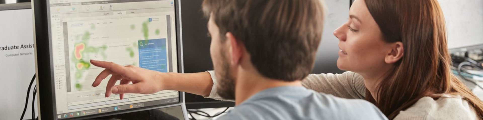 A female teacher points at a computer screen while sat next to a male student.