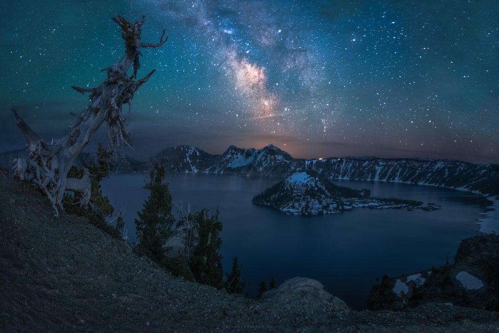 A mountain sits in a blue lake under a blue starry sky.