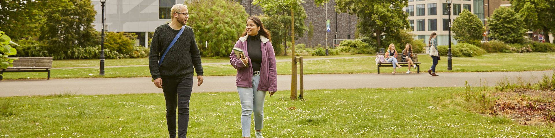 Two students walking through the park with the sports complex in the background
