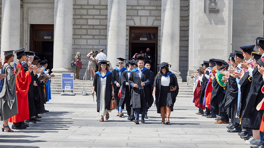 Graduates leaving Southampton Guildhall as people stand in lines either side of them and clap.