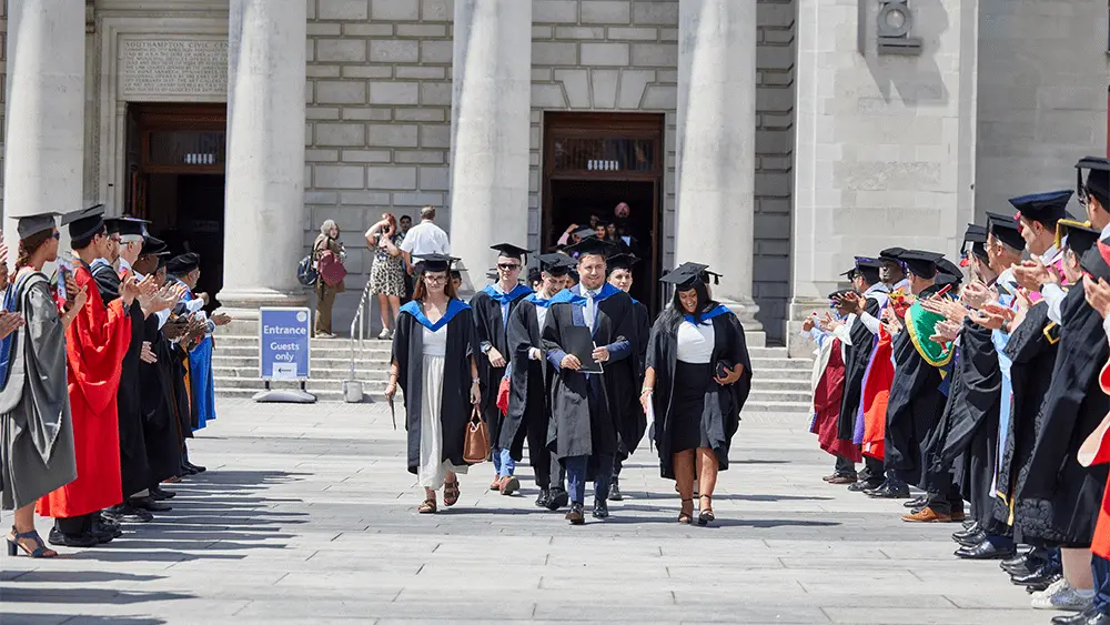Graduates leaving Southampton Guildhall as people stand in lines either side of them and clap.