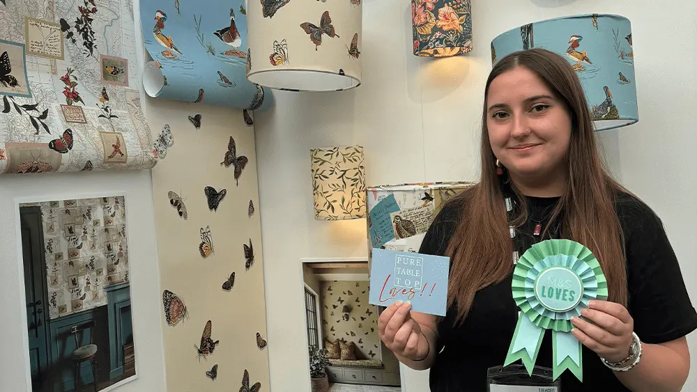 A Solent University graduate standing in front of her wallpaper and surface designs at the New Designers event.