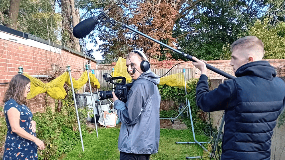 Two students holding camera equipment, filming a woman in her garden.