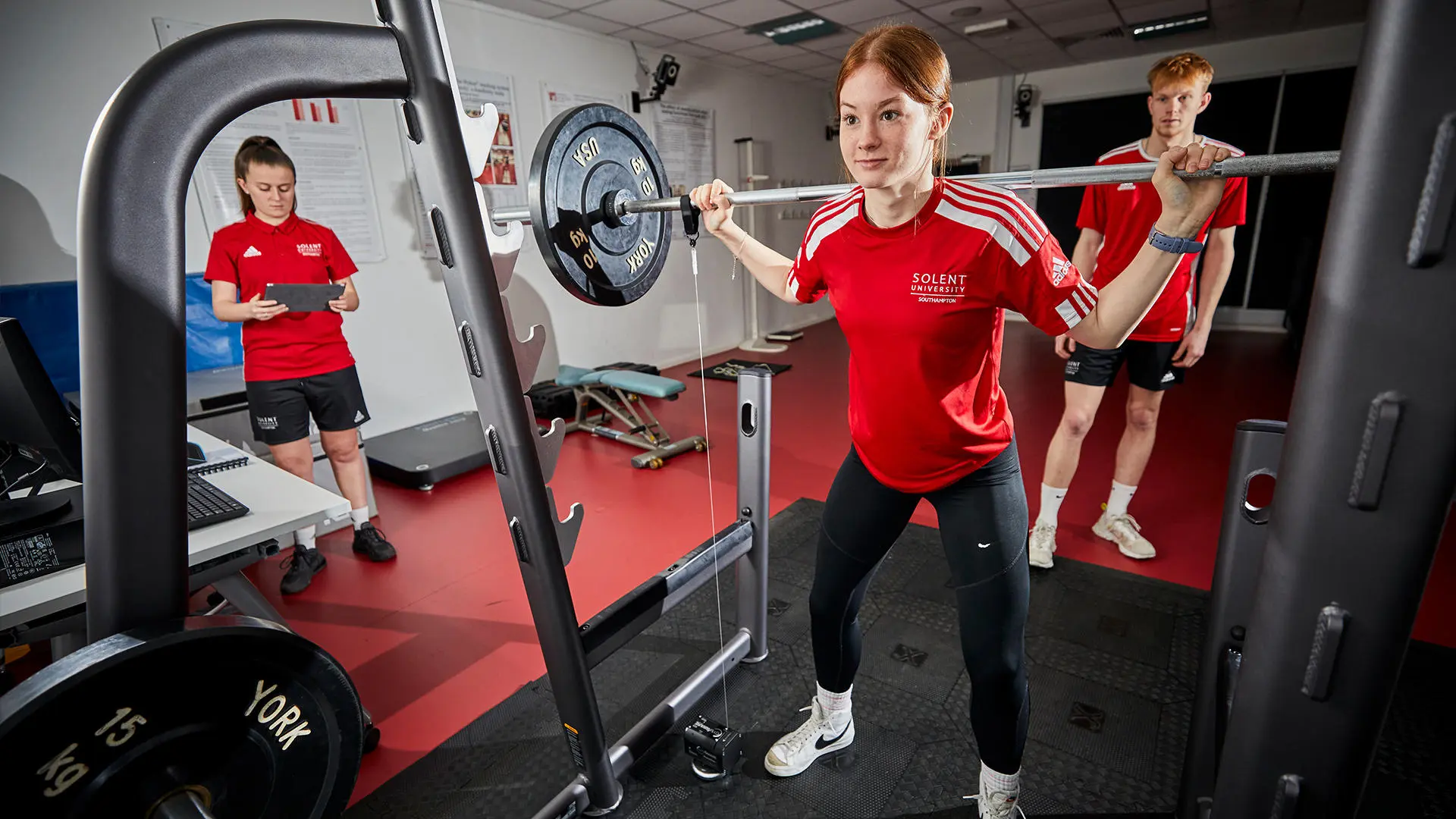 Image of student working in biomechanics lab - a student is doing a barbell back squat whilst two other students analyse the movements on a computer. 