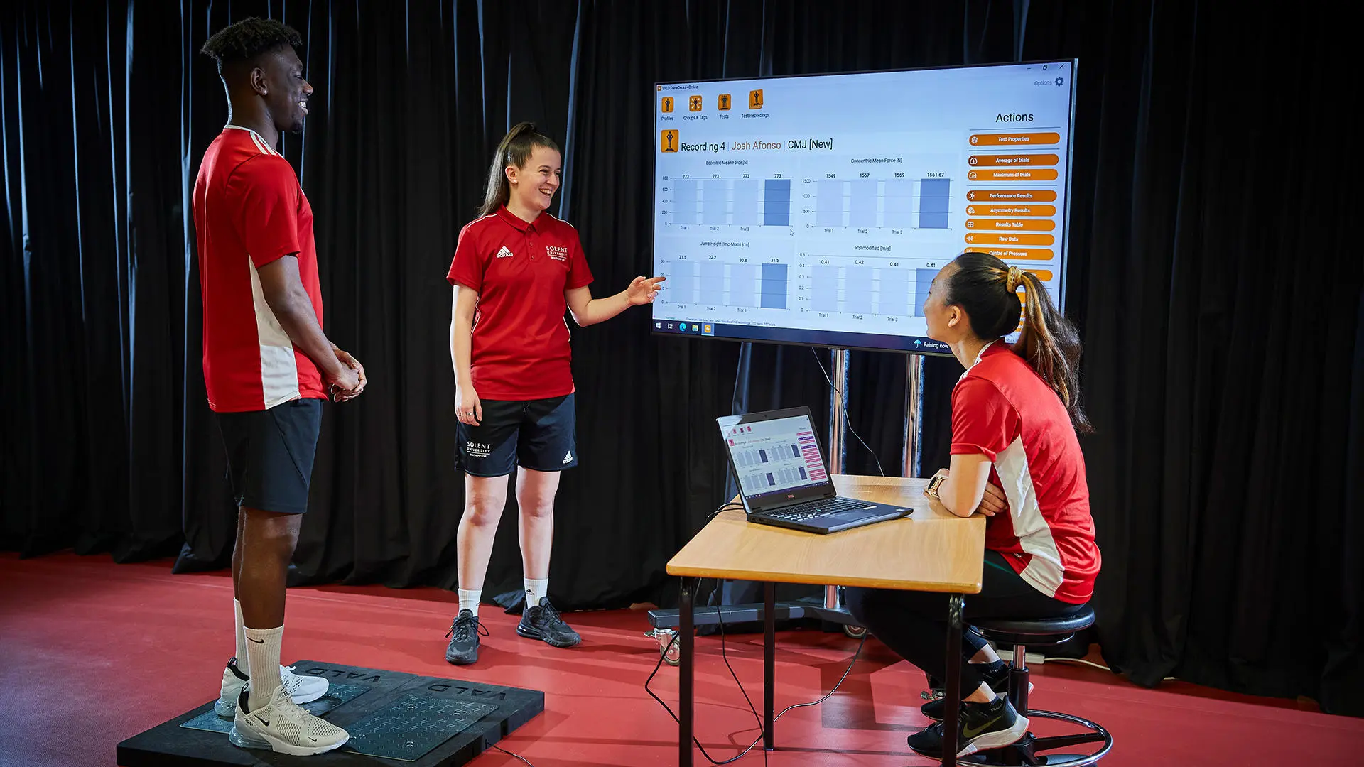 Image of student working in biomechanics lab - the three students are looking at the results on a computer and large screen. 