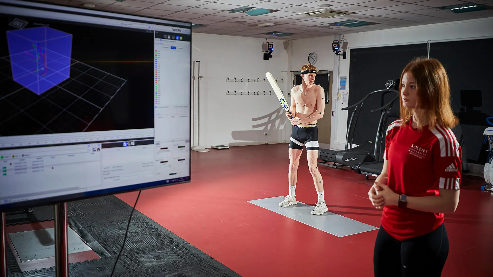 Image of student working in biomechanics lab - a male student is holding a cricket bat and a female student is analysing the movement data on a computer.