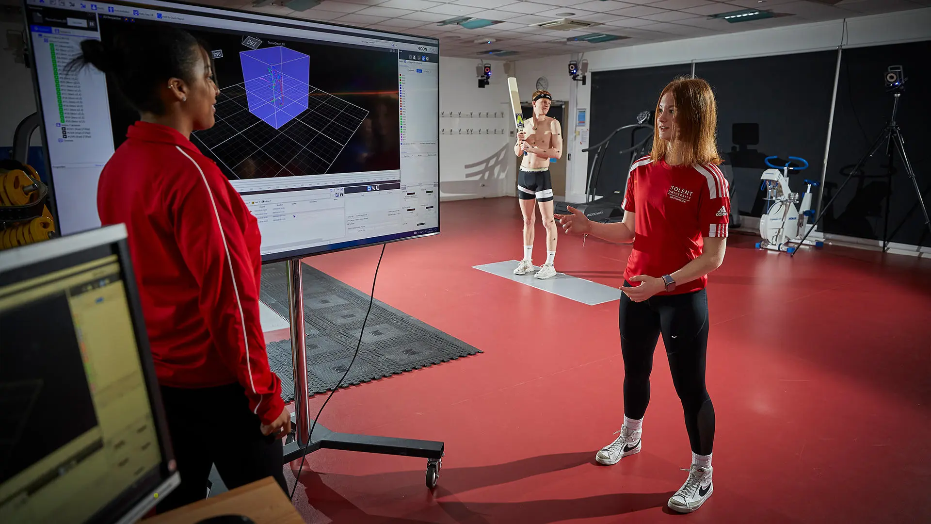 Image of student working in biomechanics lab - a male student is holding a cricket bat and two female student is analysing the movement data on a computer.