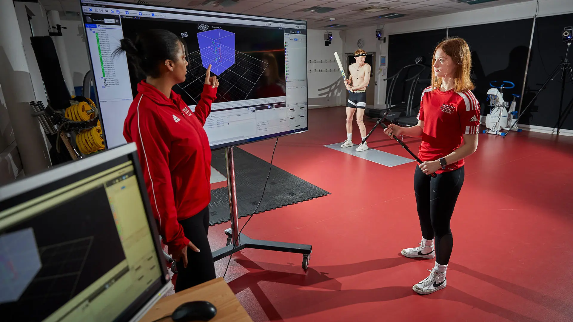 Image of student working in biomechanics lab - a male student is holding a cricket bat and two female student is analysing the movement data on a computer.