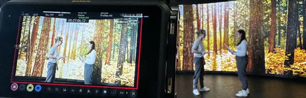 Two people stood in front of a digital screen with a close up of a camera screen filming them