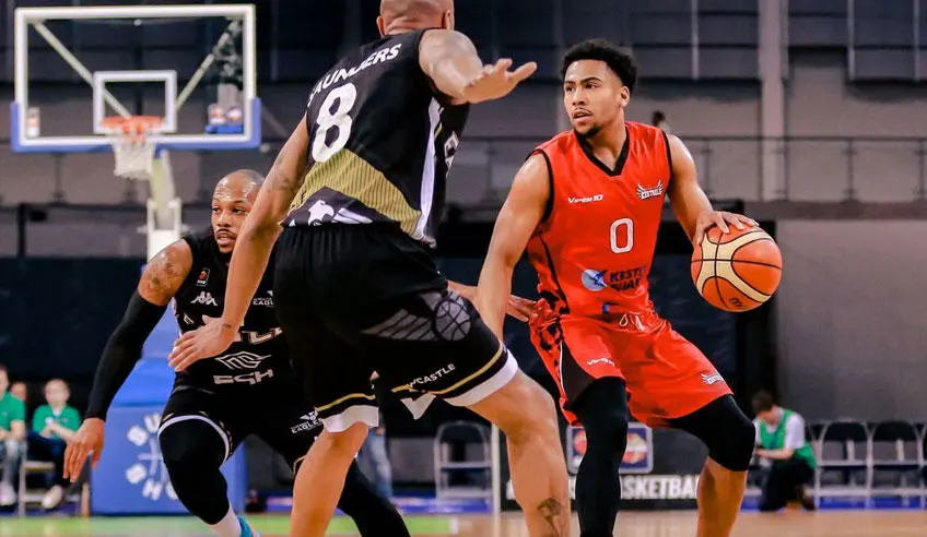 A basketball player in red with the ball in his hand going past a player in black