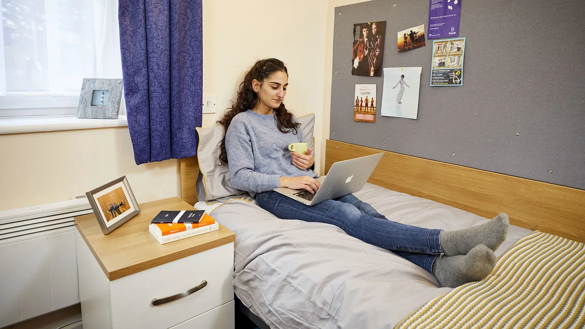 A female student sat on her bed in student accommodation looking at a laptop and holding a mug