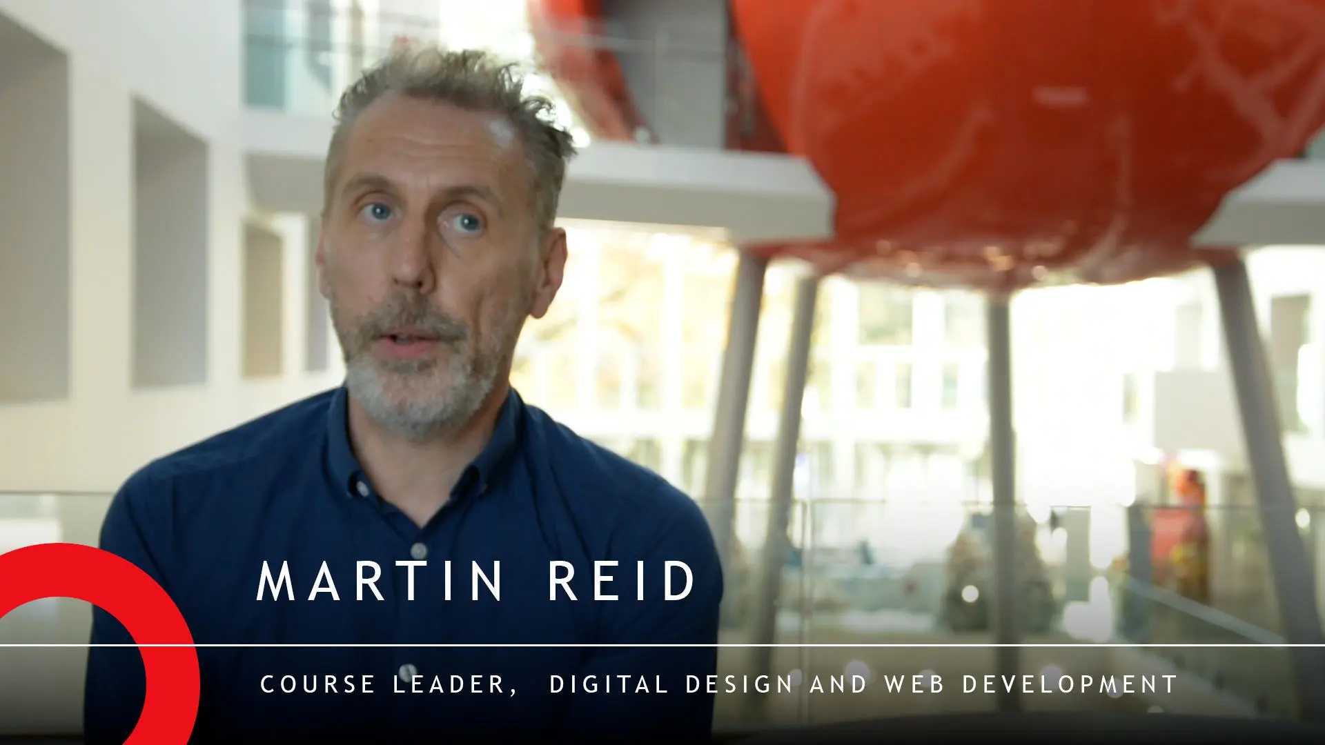 Course leader of Digital Design and Web Development, Martin Reid is sat in The Spark building at Solent University, with the red Pod out of focus in the background. His name and job description are displayed in white text at the bottom of the screen with a red semi circle bottom left of the screen.