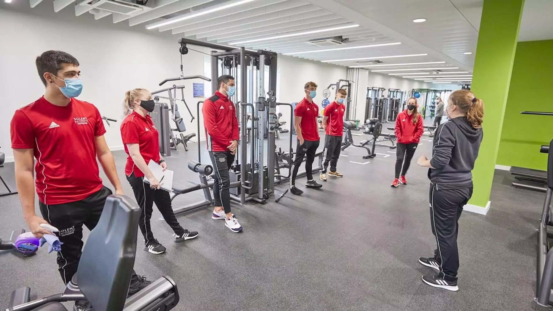 BSc (Hons) Sport Management students stood in Solent University sports uniform, wearing face masks, being taught by a lecturer in the gym at Solent's Sport Complex.