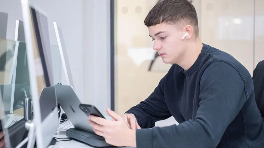 A male student sat in front of a desktop computer looking at a smartphone