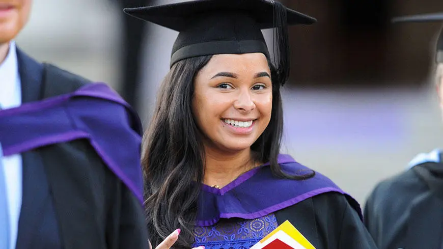 A female graduate wearing a cap and gown and smiling at the camera