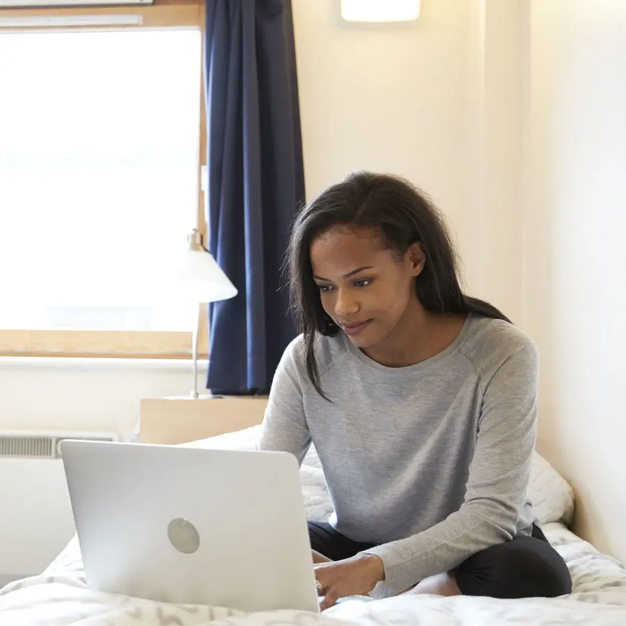 Female student sat on a bed looking at a laptop