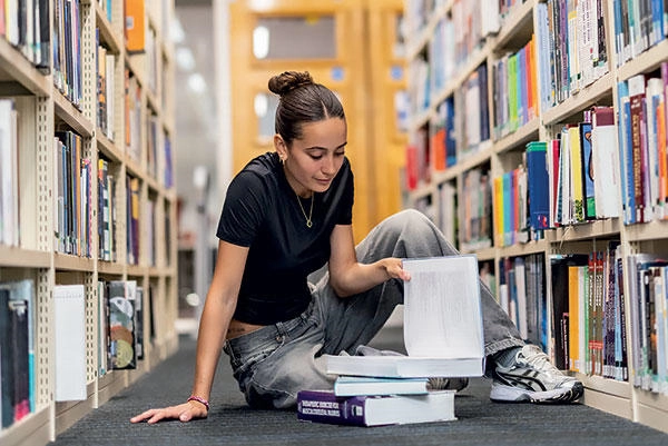 A female student sitting on the floor of a library, reading books