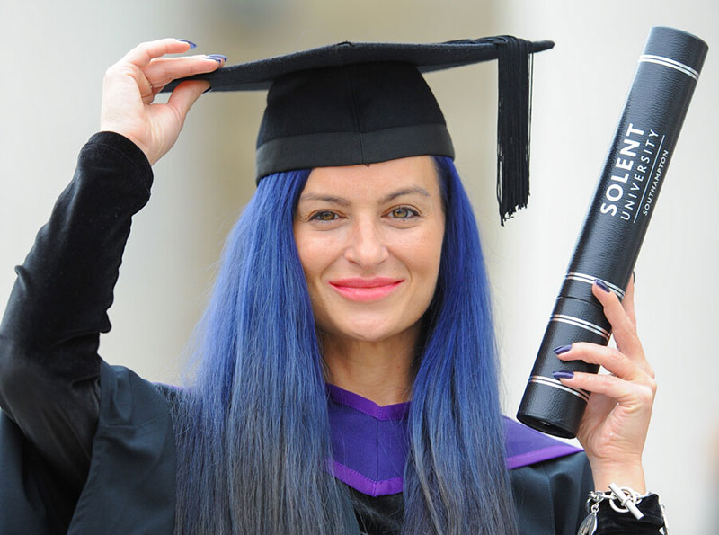 A graduate with blue hair holding her degree certificate
