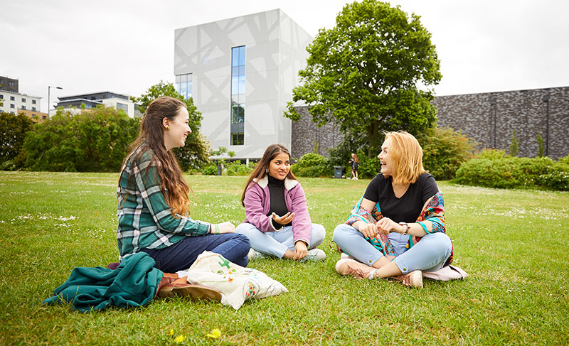 Three female students sat on the grass in the park laughing