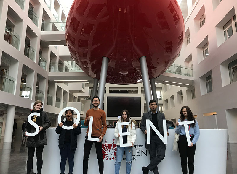 Solent's team of international alumni ambassadors standing in front of the pod holding a letters to spell Solent