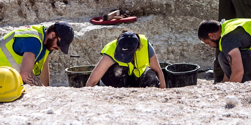 Three people in hi-vis tabards digging on an archaeological site