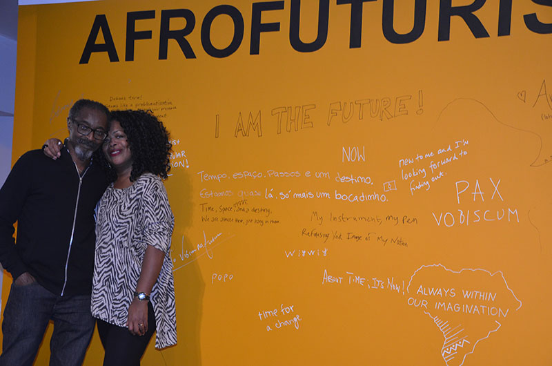 Don John and friend in front of the yellow wall that has graffiti on it