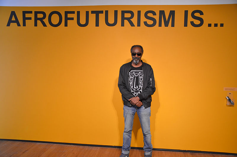 Don John in front of a yellow wall with the words 'Afrofuturism is...' on it