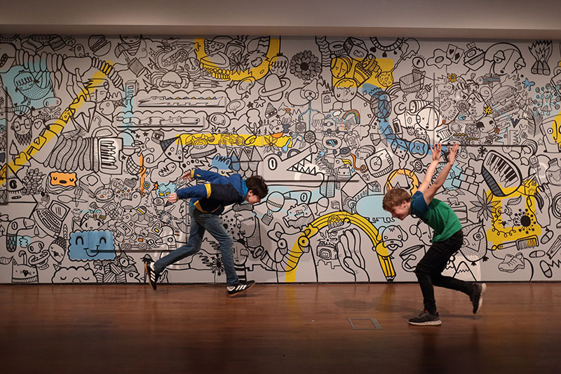 Two children running in front of a Kev Munday mural